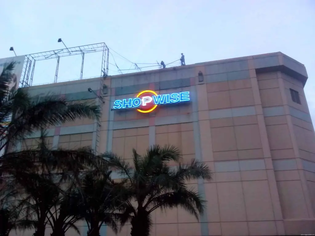 Neon SHOPWISE signage on large beige building in Quezon City with palm trees and workers on roof, by M&G Global Ads.