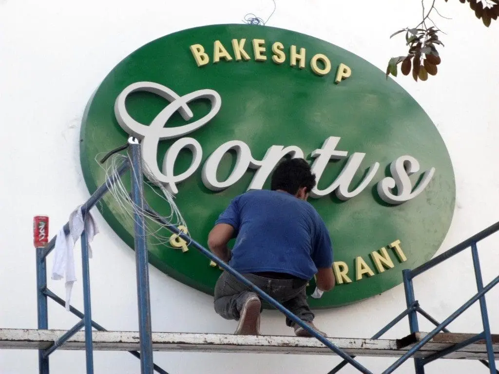 Acrylic signage for Cortys Bakeshop & Restaurant installation in Quezon City by M&G Global Ads. Worker on scaffolding, tools visible.