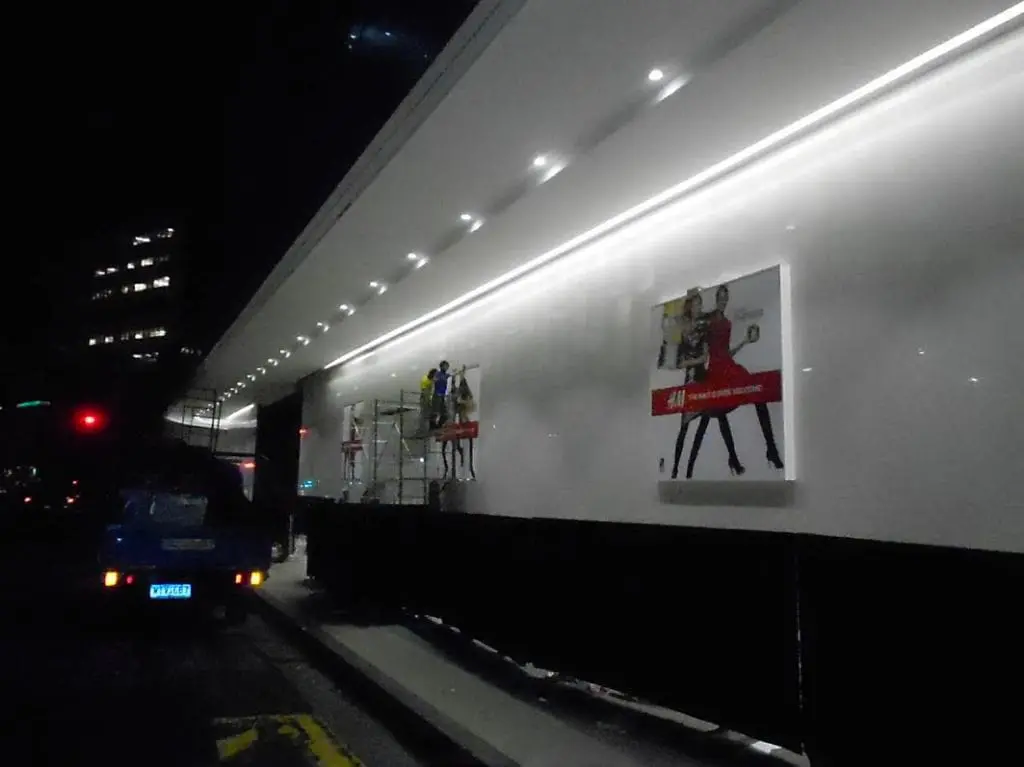 Workers install illuminated building signage at night in Makati with a blue car nearby, by M&G Global Ads.
