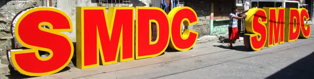 Acrylic SMDC and SMDS signage in bold red and yellow on a sunny Quezon City street, with passerby and dog—by M&G Global Ads.