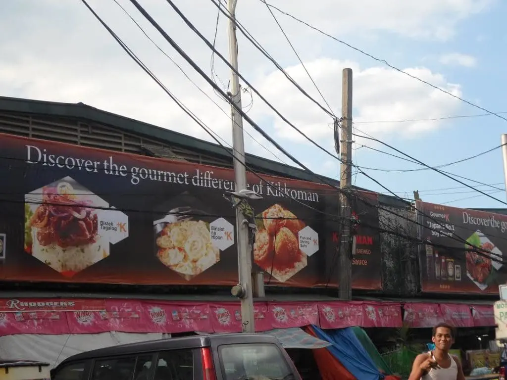 Kikkoman billboard signage on a Makati building above street stalls with power lines and a person eating ice cream. By M&G Global Ads.