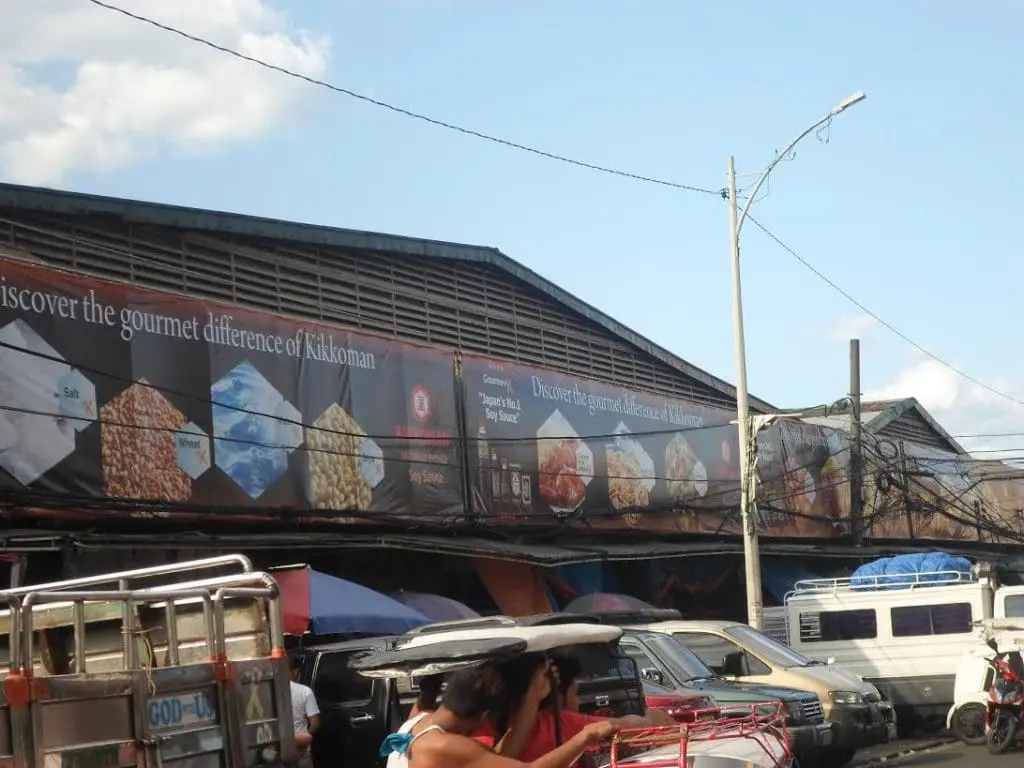 Outdoor Kikkoman banner signage at busy market scene in Quezon City, showing food images and slogan, by M&G Global Ads.