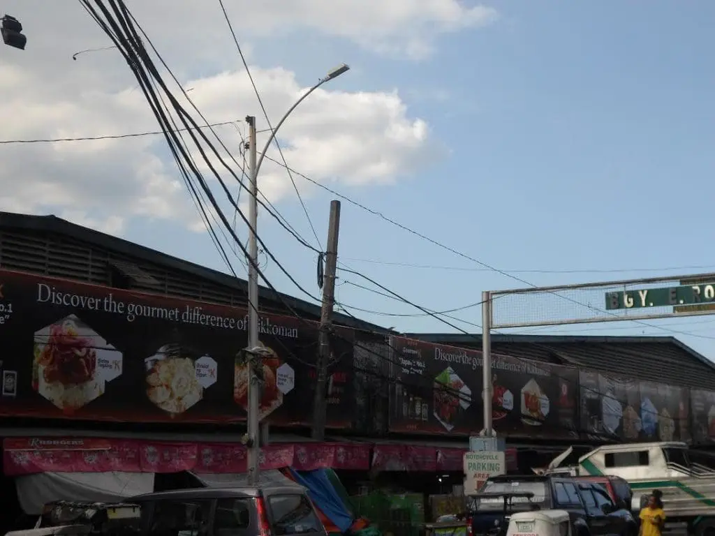 Gourmet food billboard and outdoor signage on dark building in Makati, tangled wires, parking sign, cars—by M&G Global Ads.