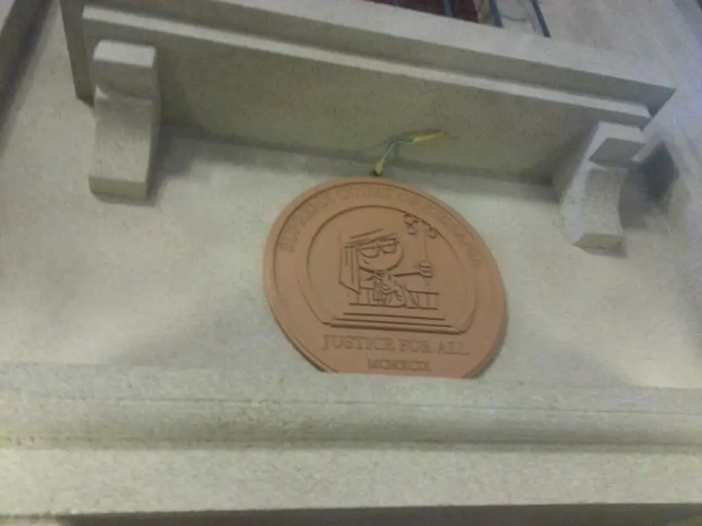 Round metallic Supreme Court office sign with cartoon judge on stone wall in Quezon City, by M&G Global Ads. Slightly blurred.