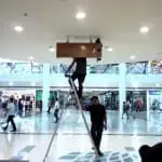 Brown acrylic indoor signage installation at a busy Quezon City mall, with workers on ladder. By M&G Global Ads.