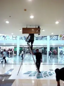 Brown acrylic indoor signage installation at a busy Quezon City mall, with workers on ladder. By M&G Global Ads.