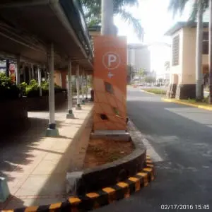 Outdoor parking signage with white P and left arrow on orange, landscaped island in Makati, palm trees, by M&G Global Ads.