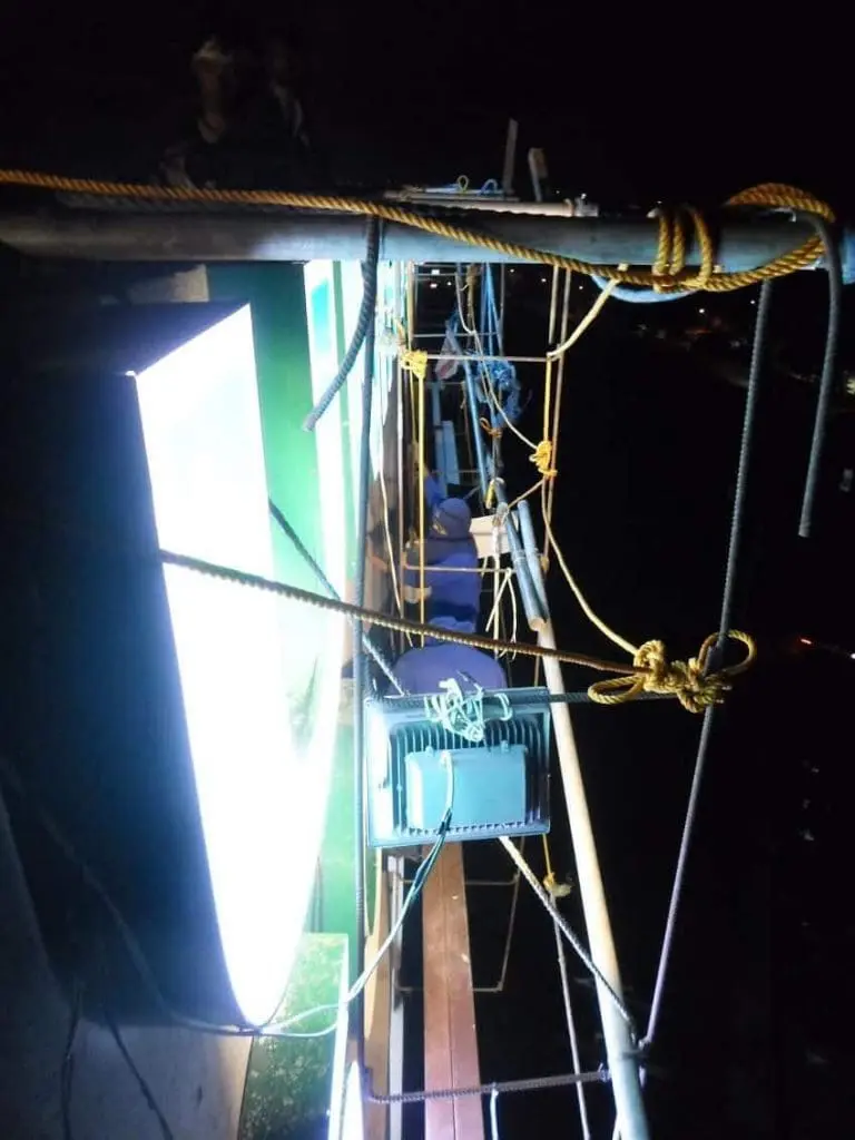 Acrylic signage repair at night with tools and ropes, worker’s legs visible, Ortigas city view by M&G Global Ads.