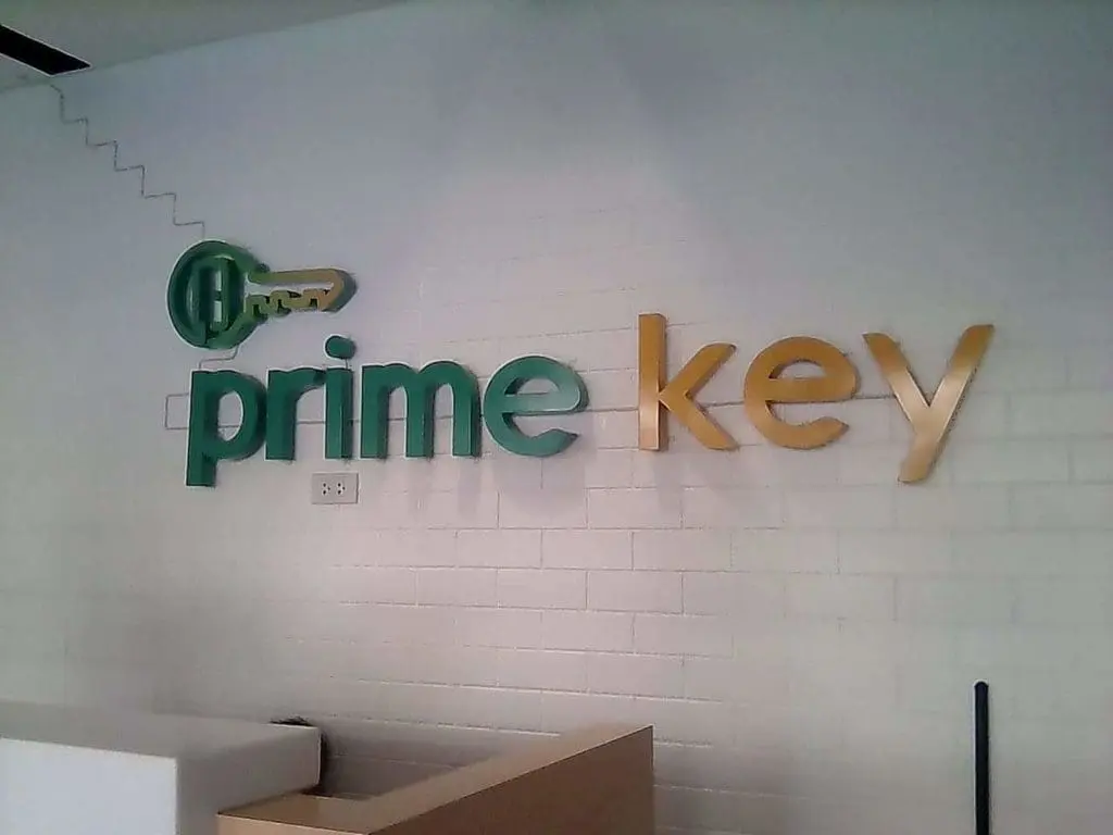 Acrylic indoor signage for Prime Key in green and gold on a white brick wall, Makati office reception by M&G Global Ads.