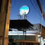 7-Eleven store signage installation Quezon City, worker on scaffolding at dusk, jeepney below, power lines. by M&G Global Ads.