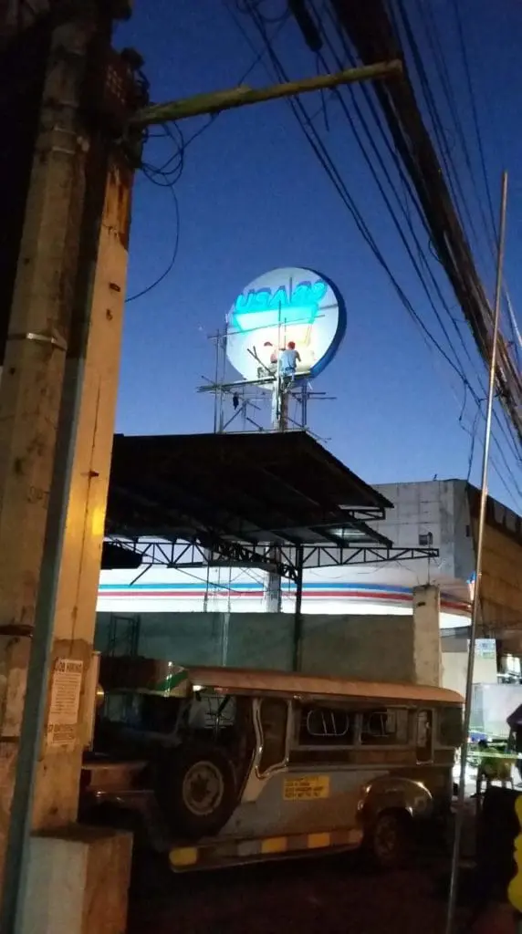 7-Eleven store signage installation Quezon City, worker on scaffolding at dusk, jeepney below, power lines. by M&G Global Ads.