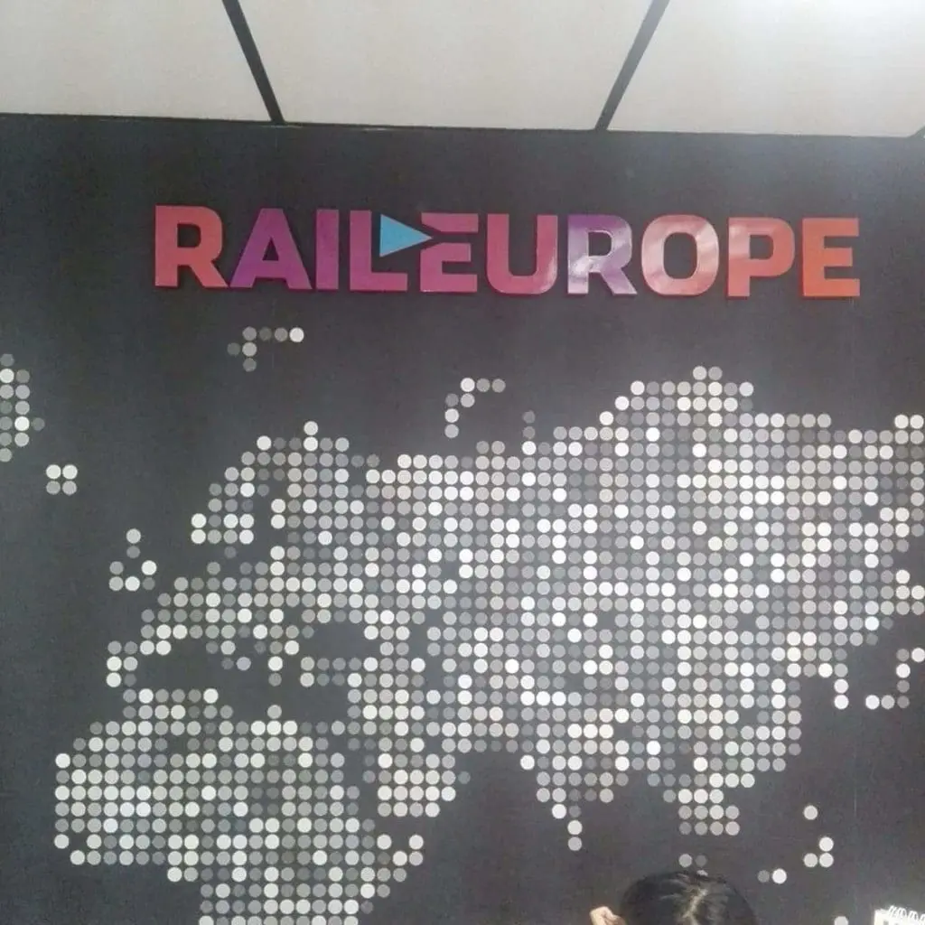 Rail Europe metallic logo signage on wall with dotted Europe map, in BGC, by M&G Global Ads. Part of a persons head visible.