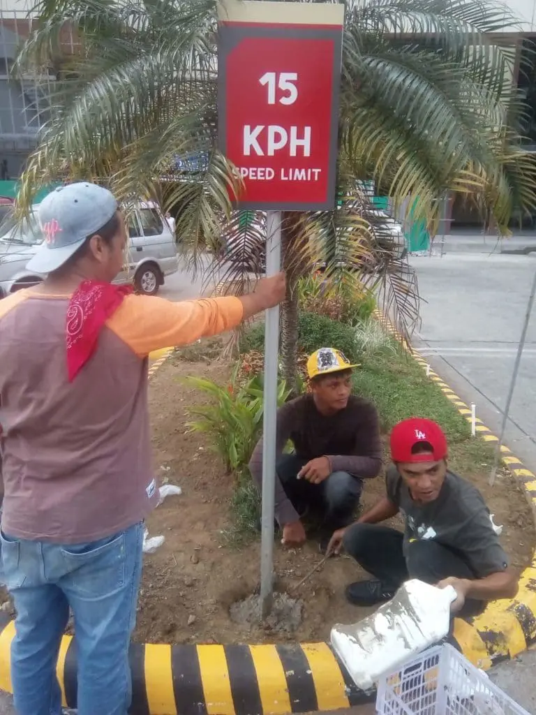 Building speed limit signage installation, 15 KPH, landscaped median in Quezon City by M&G Global Ads. Three men at work.