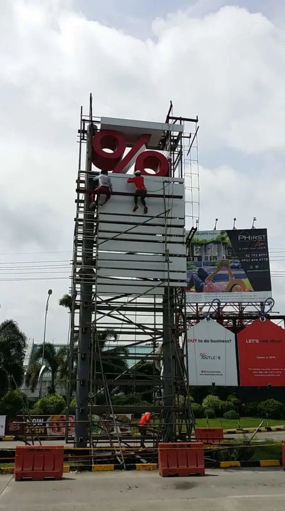 Workers installing a roadside billboard sign with red percent symbol in Quezon City, orange barriers visible, by M&G Global Ads.