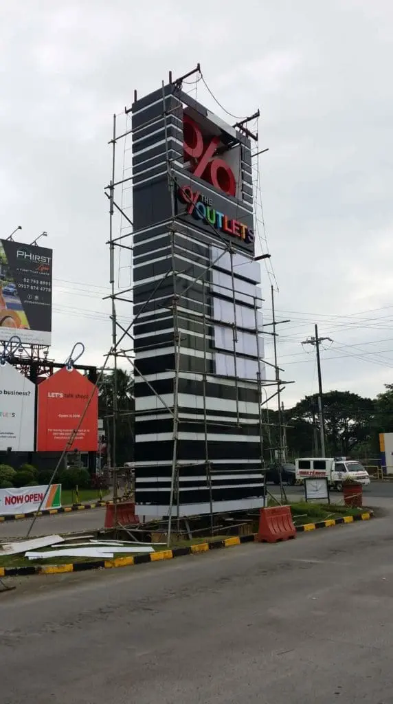 Roadside pylon signage for THE OUTLETS under construction in Mandaluyong, traffic barriers, cars; by M&G Global Ads.