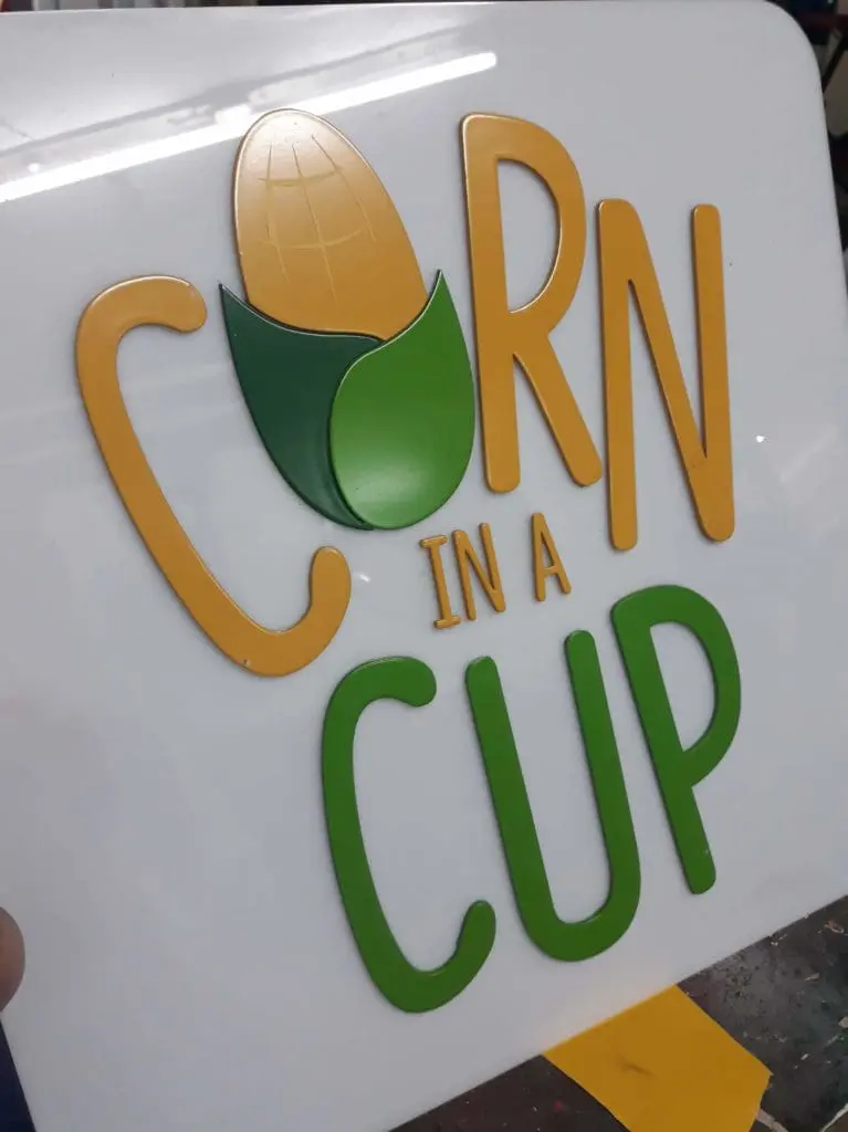Acrylic signage for CORN IN A CUP with corn cob art in Ortigas, yellow and green letters on white by M&G Global Ads.