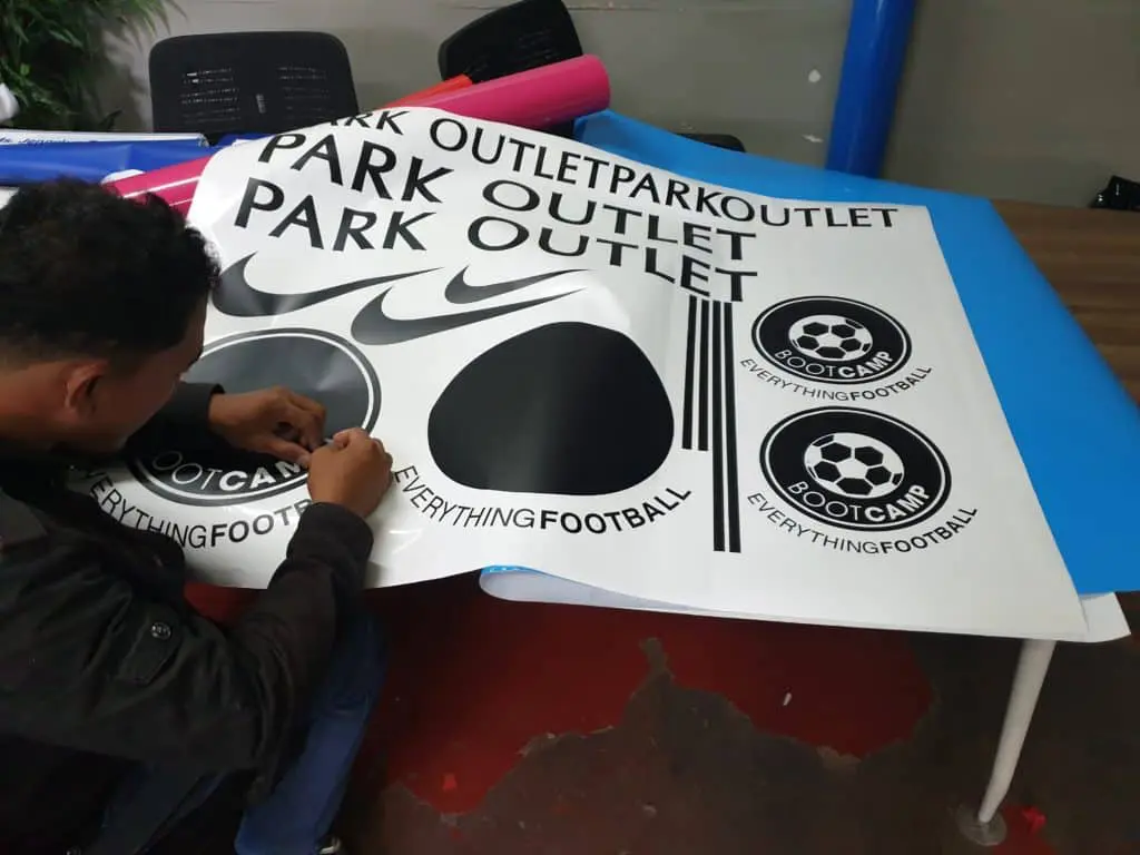 Outdoor signage printing for PARK OUTLET with football logos in Pasig, vinyl rolls visible on table by M&G Global Ads.