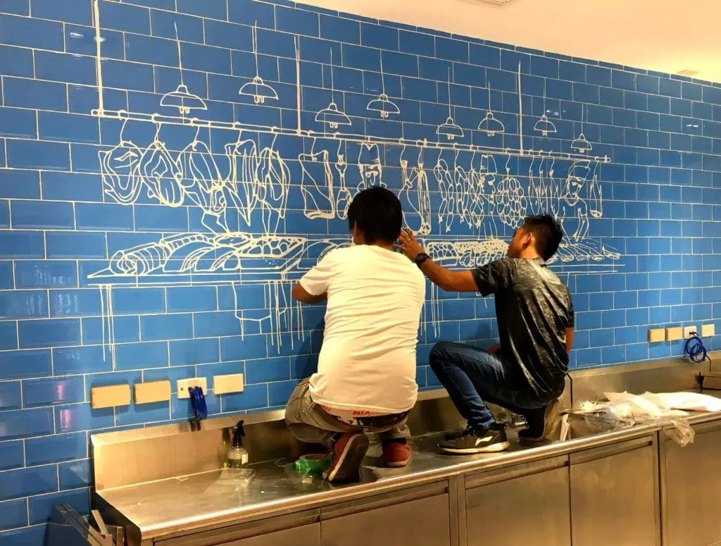 Artists painting a meat mural on blue tiles under bold LED store signage in Makati, stainless counter; by M&G Global Ads.