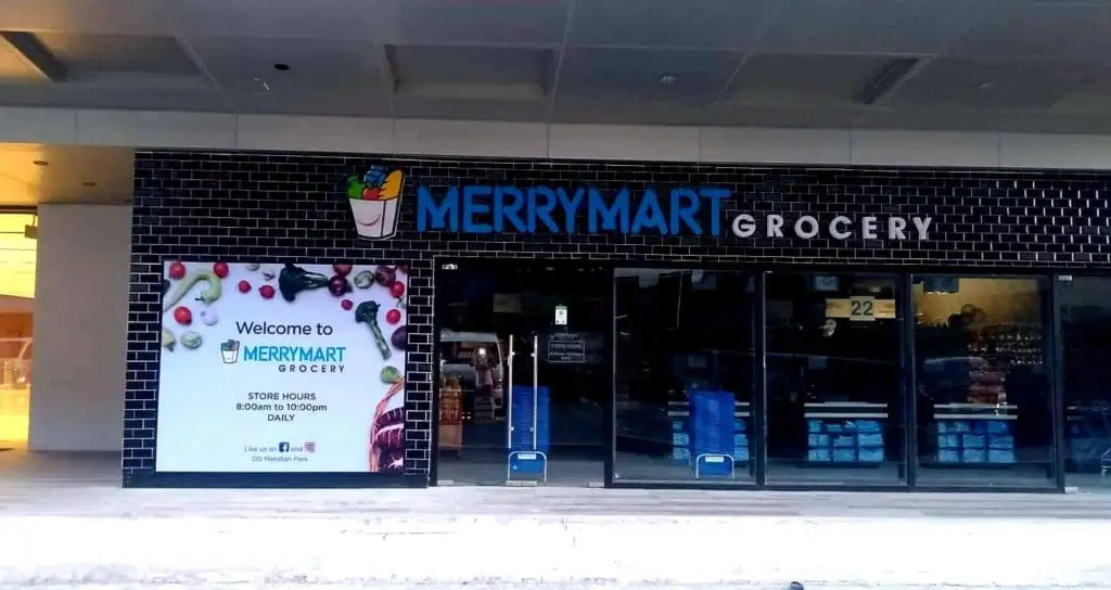 Metallic storefront signage for MERRYMART Grocery in Quezon City, showing store hours by glass doors. By M&G Global Ads.