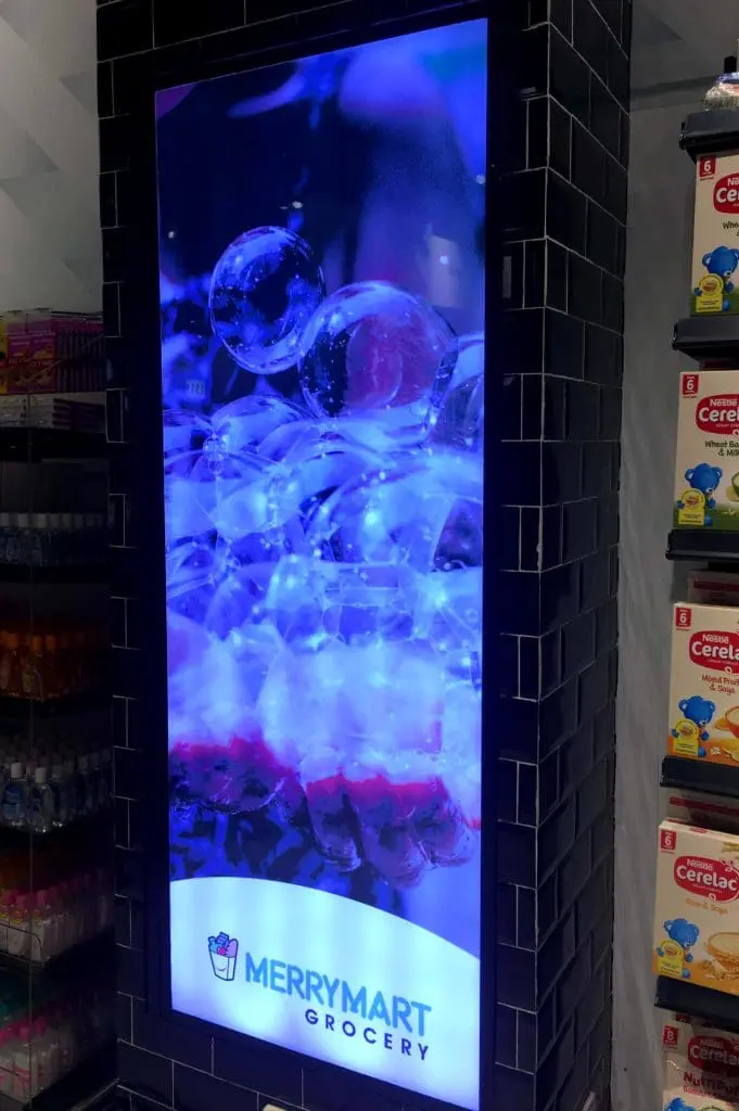 LED signage screen for MerryMart Grocery in Quezon City displays logo and abstract bubbles, store shelves beside it. By M&G Global Ads.