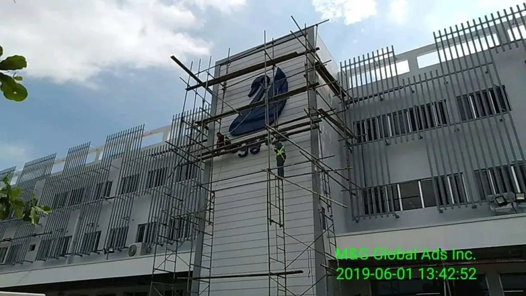 Acrylic signage installation on modern building, Quezon City project, workers on scaffolding under cloudy sky by M&G Global Ads.