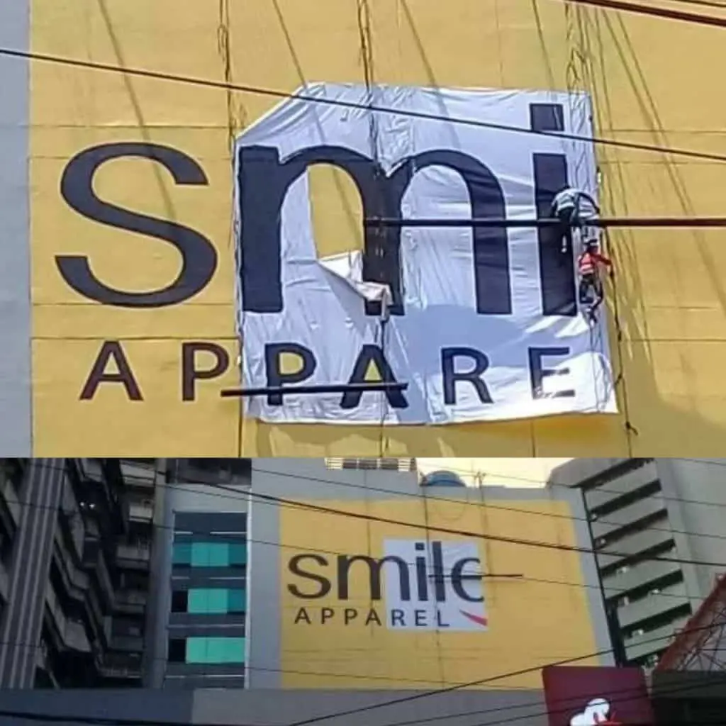 Acrylic building signage for Smile APPAREL in Makati, worker partially covers text; cityscape view. By M&G Global Ads.