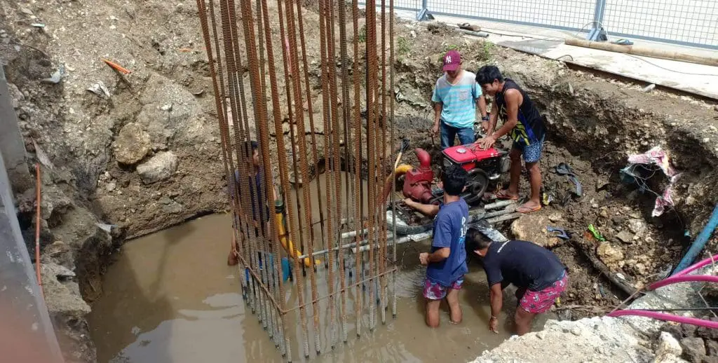 Construction workers in muddy pit with reinforced steel, operating machinery and billboard signage, Quezon City, by M&G Global Ads.