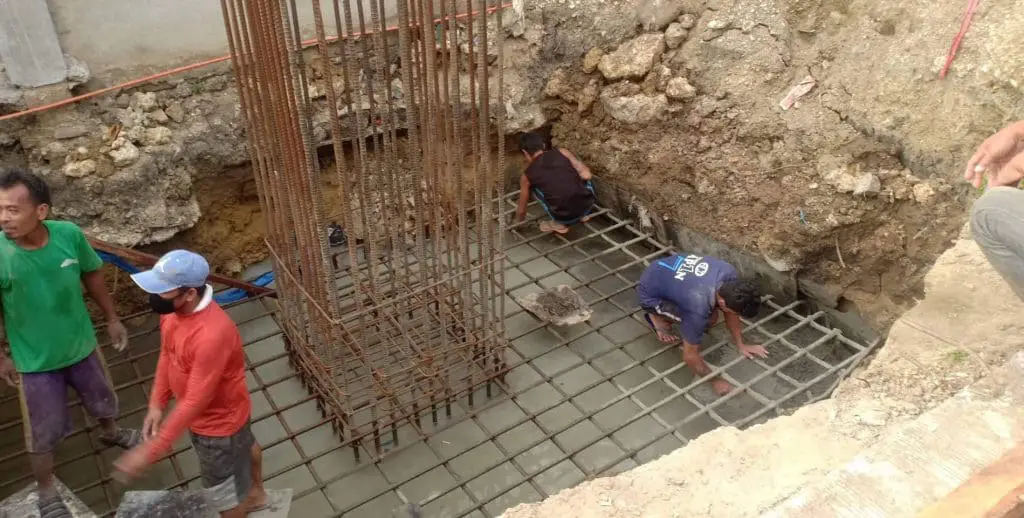 Four construction workers build a foundation in an excavated pit with outdoor tarpaulin signage in Quezon City by M&G Global Ads.