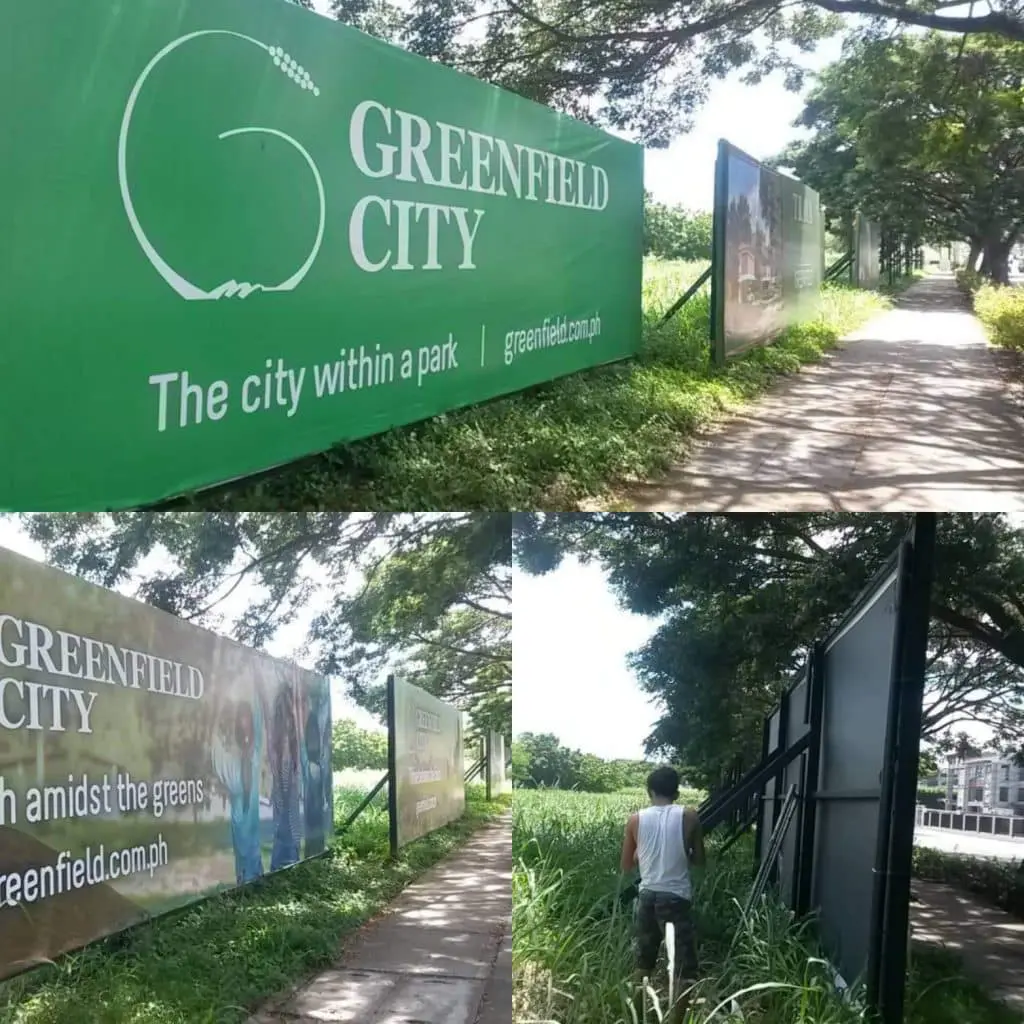 Greenfield City outdoor billboard signage in Quezon City with trees and sidewalk; person by sign and tall grass. By M&G Global Ads.