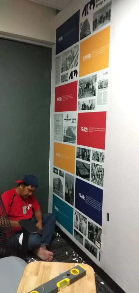 Acrylic timeline wall signage with historical photos and dates, installed in Makati by M&G Global Ads. Man, tools, chair visible.