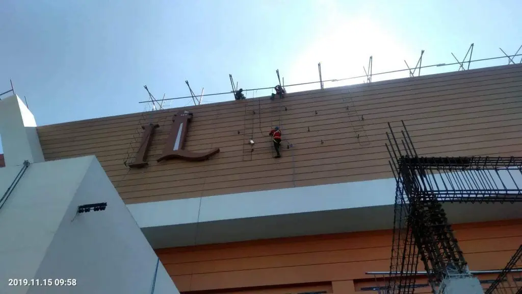 Workers installing metallic building signage on tall exterior wall in BGC, under blue sky. By M&G Global Ads.