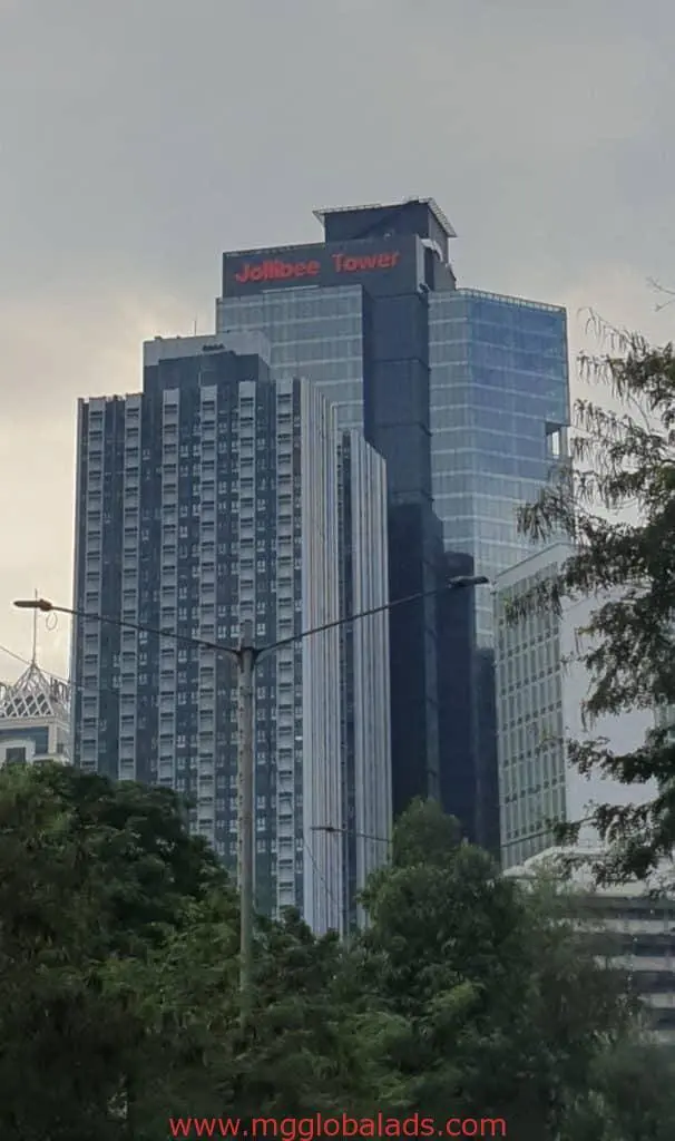 Jollibee Tower glass skyscraper with illuminated building signage in Ortigas stands tall among trees and cityscape, by M&G Global Ads.