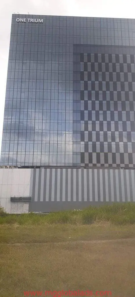 Metallic building signage ONE TRIUM on glass tower in Ortigas by M&G Global Ads; checkerboard windows, grass, cloudy sky.