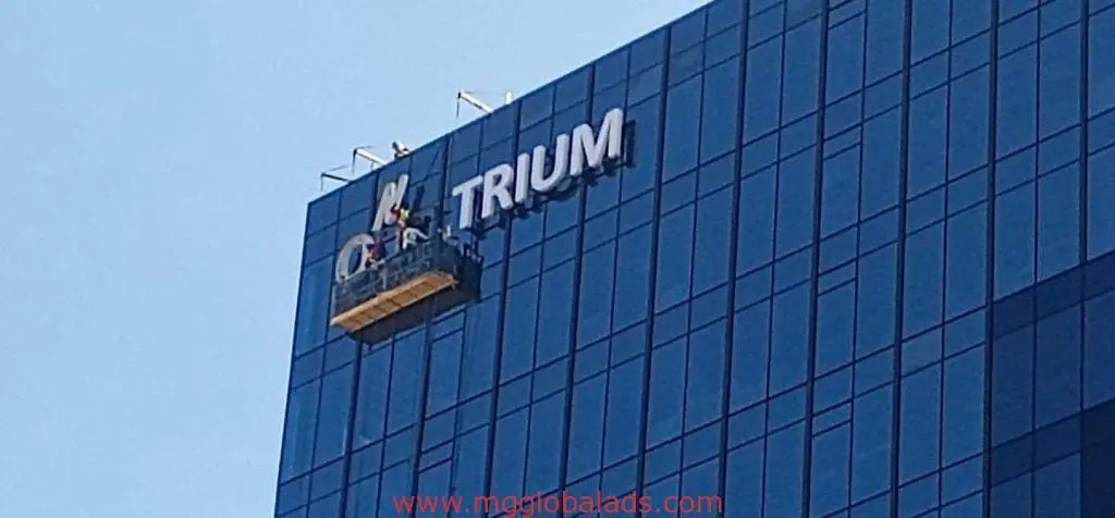 Workers install outdoor building signage for TRIUM on a glass office tower in Makati, some letters missing, by M&G Global Ads.