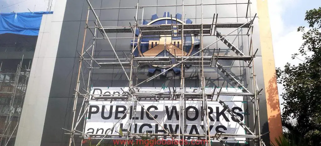 PUBLIC WORKS AND HIGHWAYS outdoor building signage in Makati with scaffolding, logo, and blue sky by M&G Global Ads.