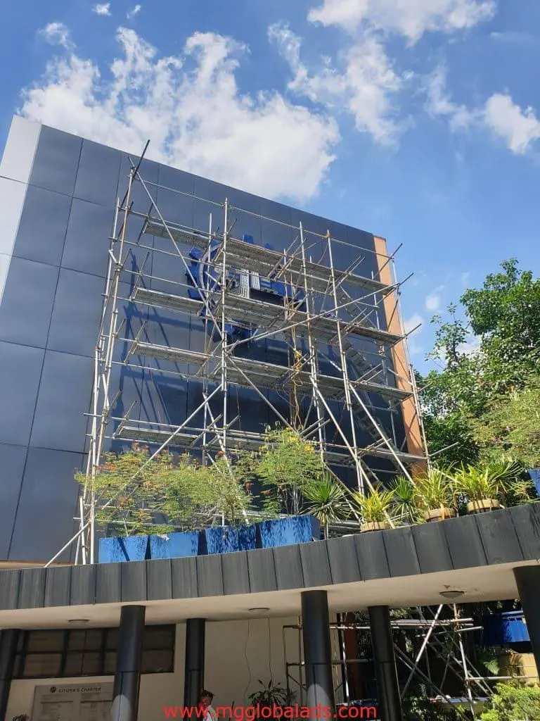 Building scaffolding with workers and outdoor business signage in front of glass facade and plants, Ortigas by M&G Global Ads.