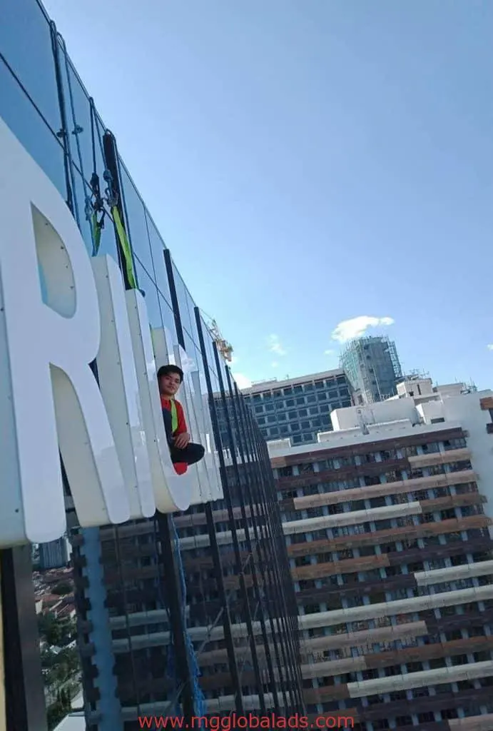 Worker hangs on ropes by acrylic signage install on glass facade in BGC, with cityscape view, by M&G Global Ads.
