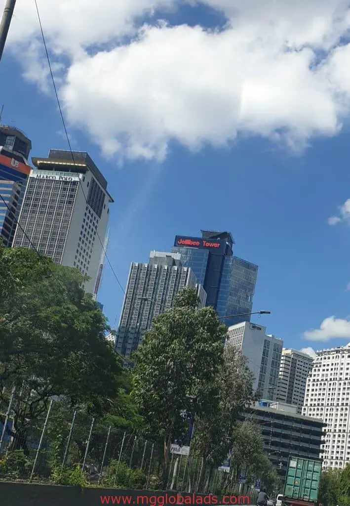 Jollibee Tower skyscraper with outdoor billboard signage in Ortigas, greenery, and fence along the road by M&G Global Ads.