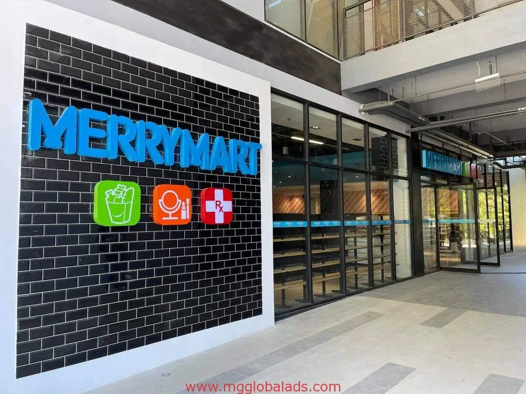 MERRYMART building signage Quezon City—blue letters, grocery, mic & Rx icons on black tile; glass shows shelves. By M&G Global Ads.