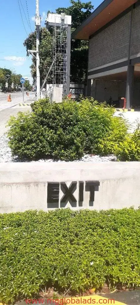 EXIT outdoor building signage on a concrete wall in Makati by M&G Global Ads, with greenery, a building, and utility pole behind.