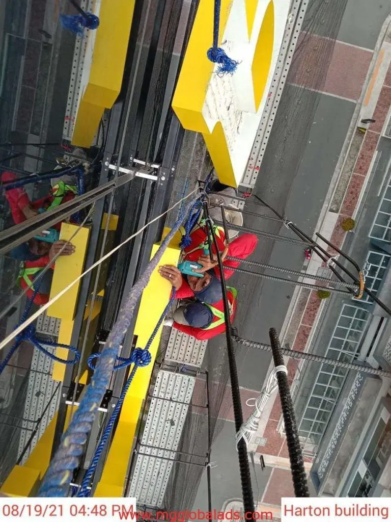 A worker cleans yellow acrylic building signage in Makati, seen from above, with street view. By M&G Global Ads.