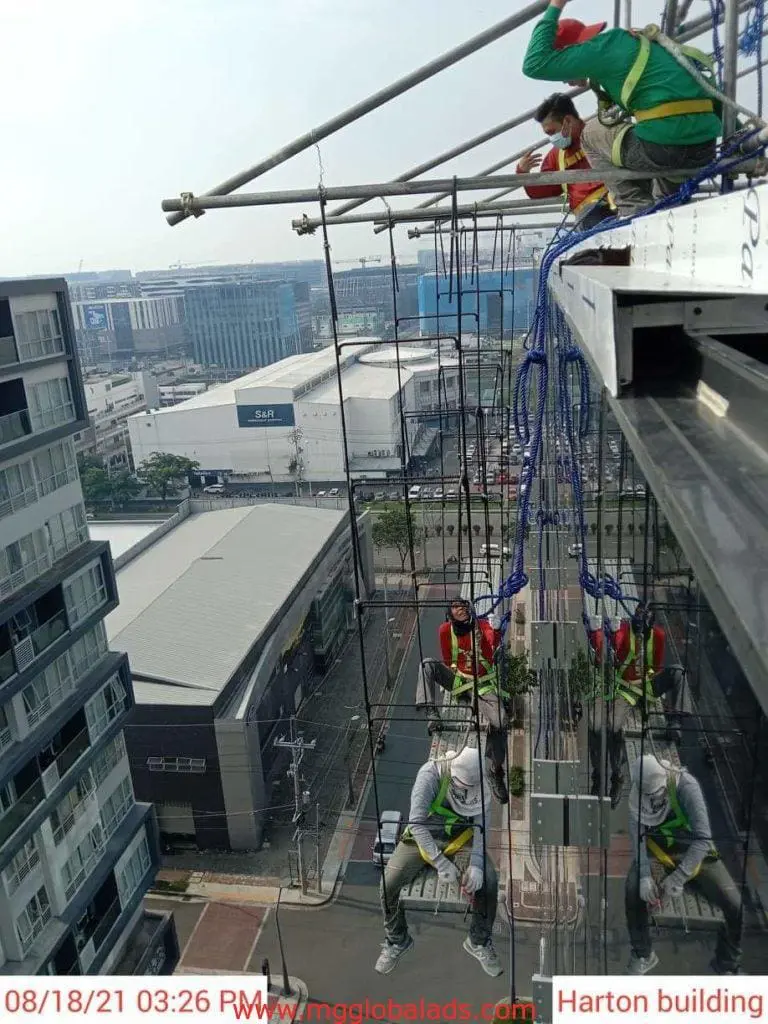 Workers in harnesses install acrylic building signage on a Makati high-rise, cityscape visible below. By M&G Global Ads.