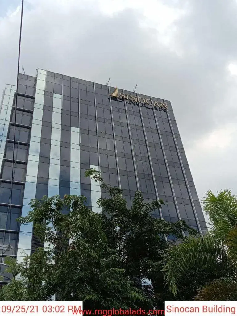 Metallic building signage for SINOCAN on a modern glass office in Makati, trees in front, cloudy sky—by M&G Global Ads.