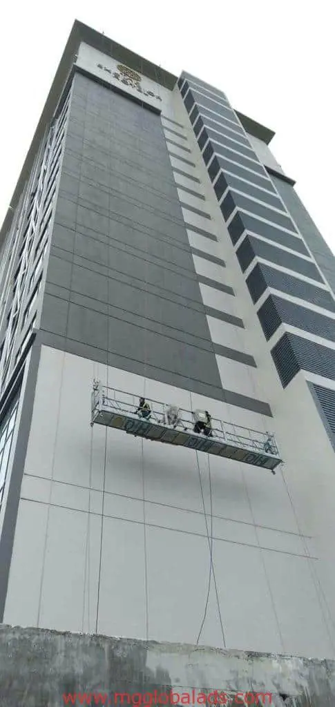 Three workers install metal building signage on a modern facade in BGC while suspended with safety ropes by M&G Global Ads.