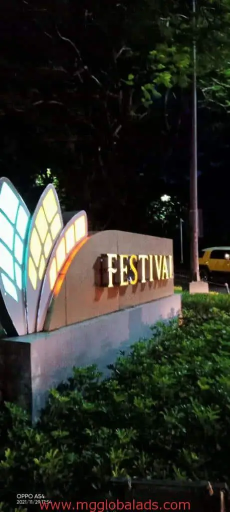Stone outdoor signage with illuminated wings and FESTIVAL in Makati, greenery and yellow vehicle at night by M&G Global Ads.
