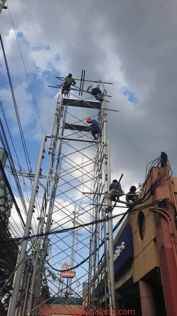 Workers installing Jollibee pylon signage on scaffold in Quezon City, with outdoor sign and electrical wires. By M&G Global Ads.