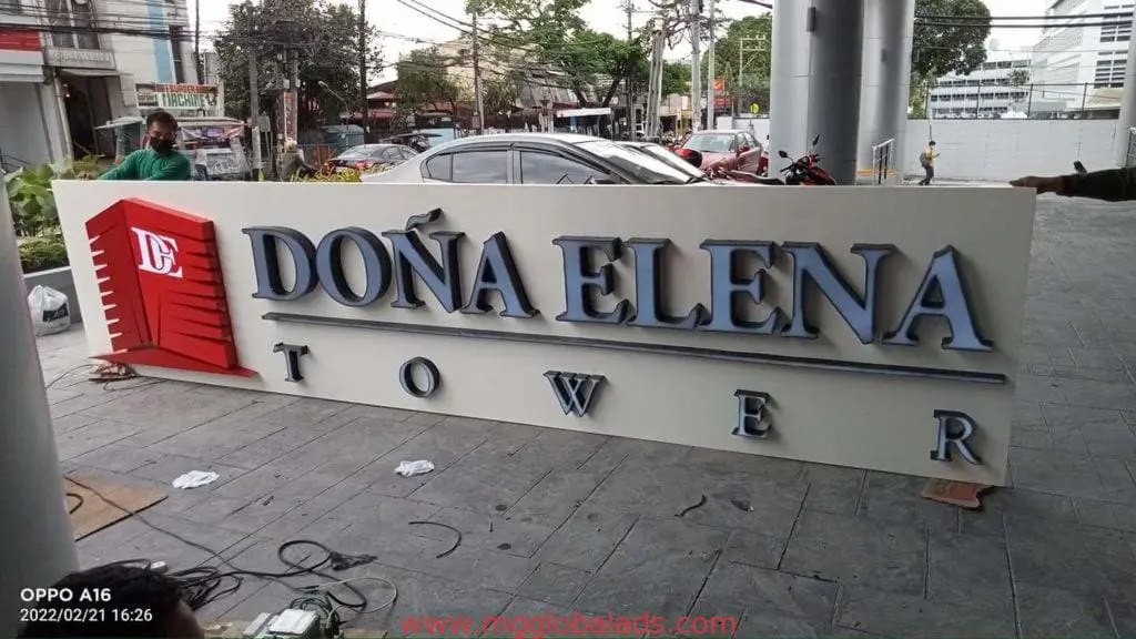 Acrylic building signage for DOÑA ELENA TOWER with red and white logo in Makati, two workers near city buildings by M&G Global Ads.