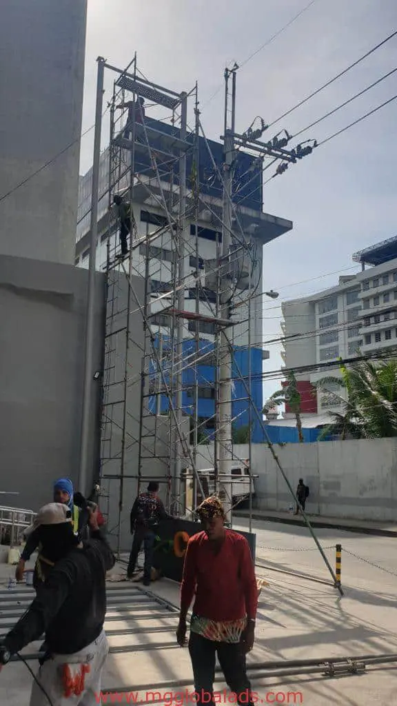 Billboard signage installation for a client in Makati, workers on scaffold, by M&G Global Ads. Urban buildings in background.