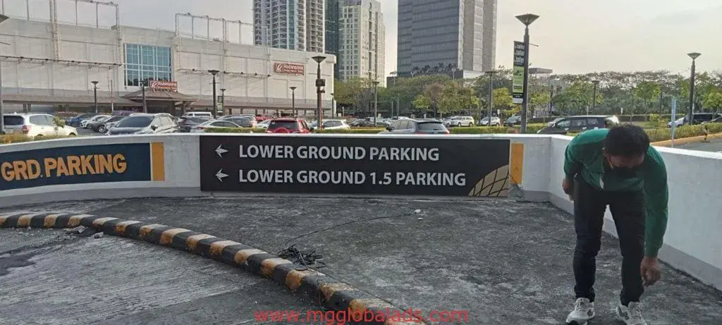 Outdoor directional parking signage at lot entrance in Quezon City, showing Lower Ground options, buildings and cars by M&G Global Ads.
