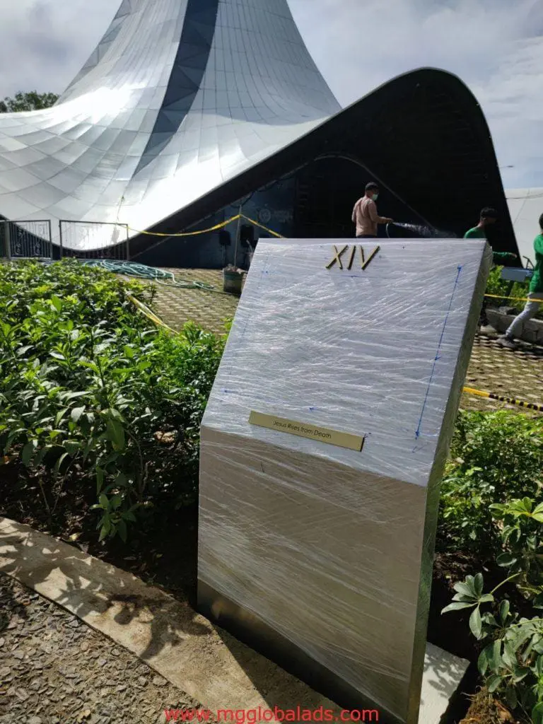 Silver metallic pylon signage XIV in Makati, wrapped in plastic near a modern building with plants and workers, by M&G Global Ads.